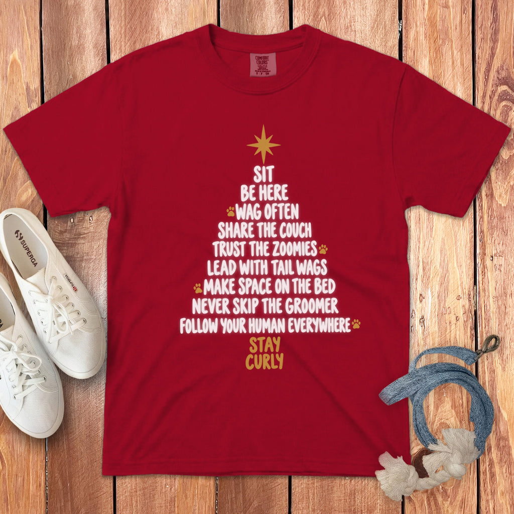 A red t-shirt printed with a dog-themed Christmas tree graphic, accompanied by white sneakers and a dog leash on a wooden background.