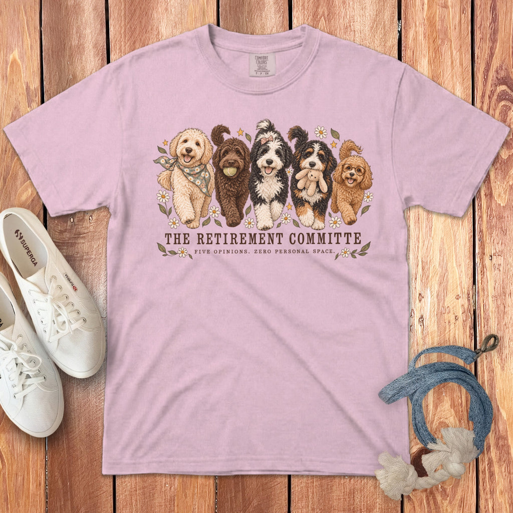 A light pink t-shirt featuring an illustration of five fluffy dogs and text reading "The Retirement Committee."