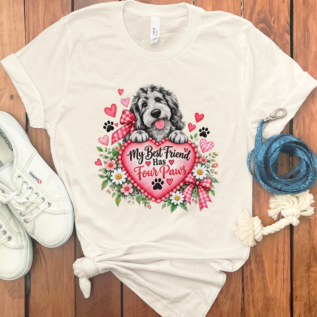 A cream-colored t-shirt with a graphic of a fluffy dog resting on a pink heart surrounded by flowers and the text My Best Friend Has Four Paws, laid flat on a wooden surface with white sneakers and a blue leash.