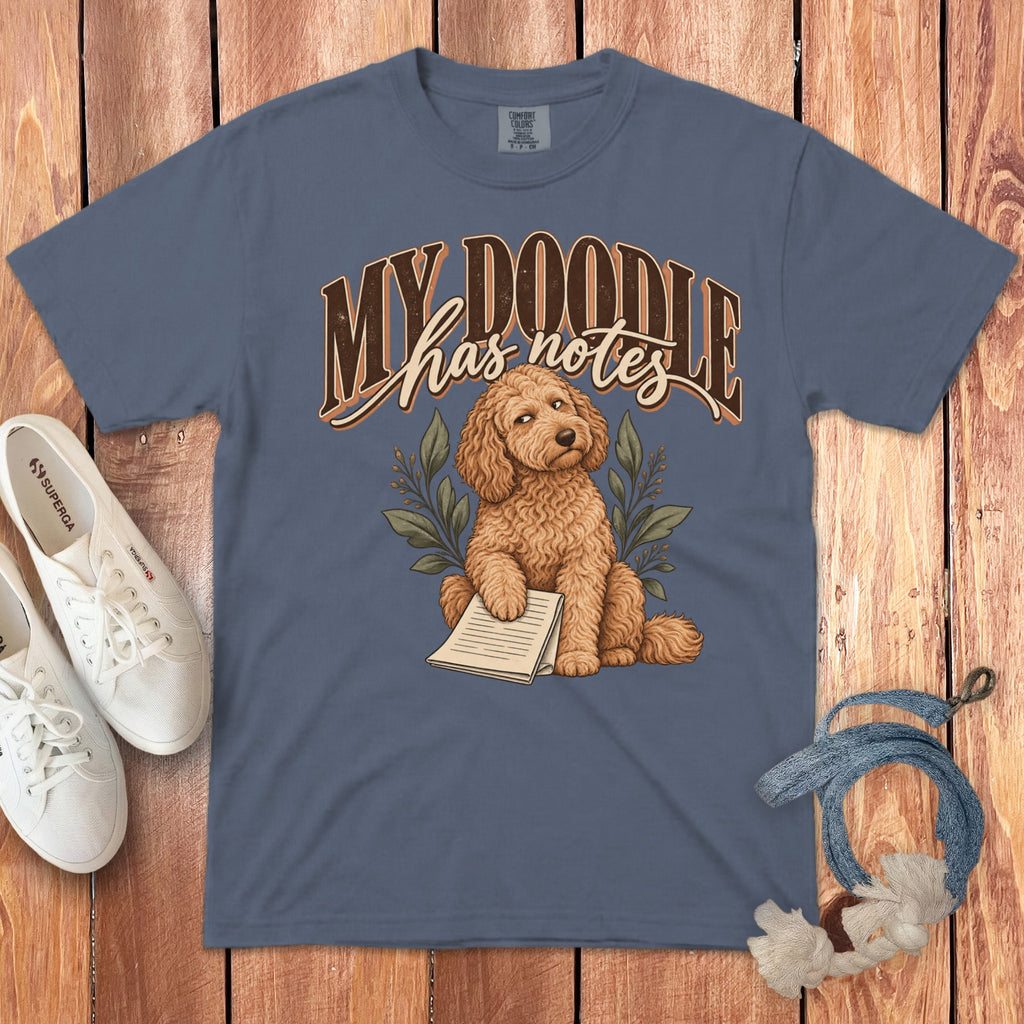 Slate blue t-shirt featuring a cartoon doodle dog holding notes, surrounded by white sneakers and a leash.