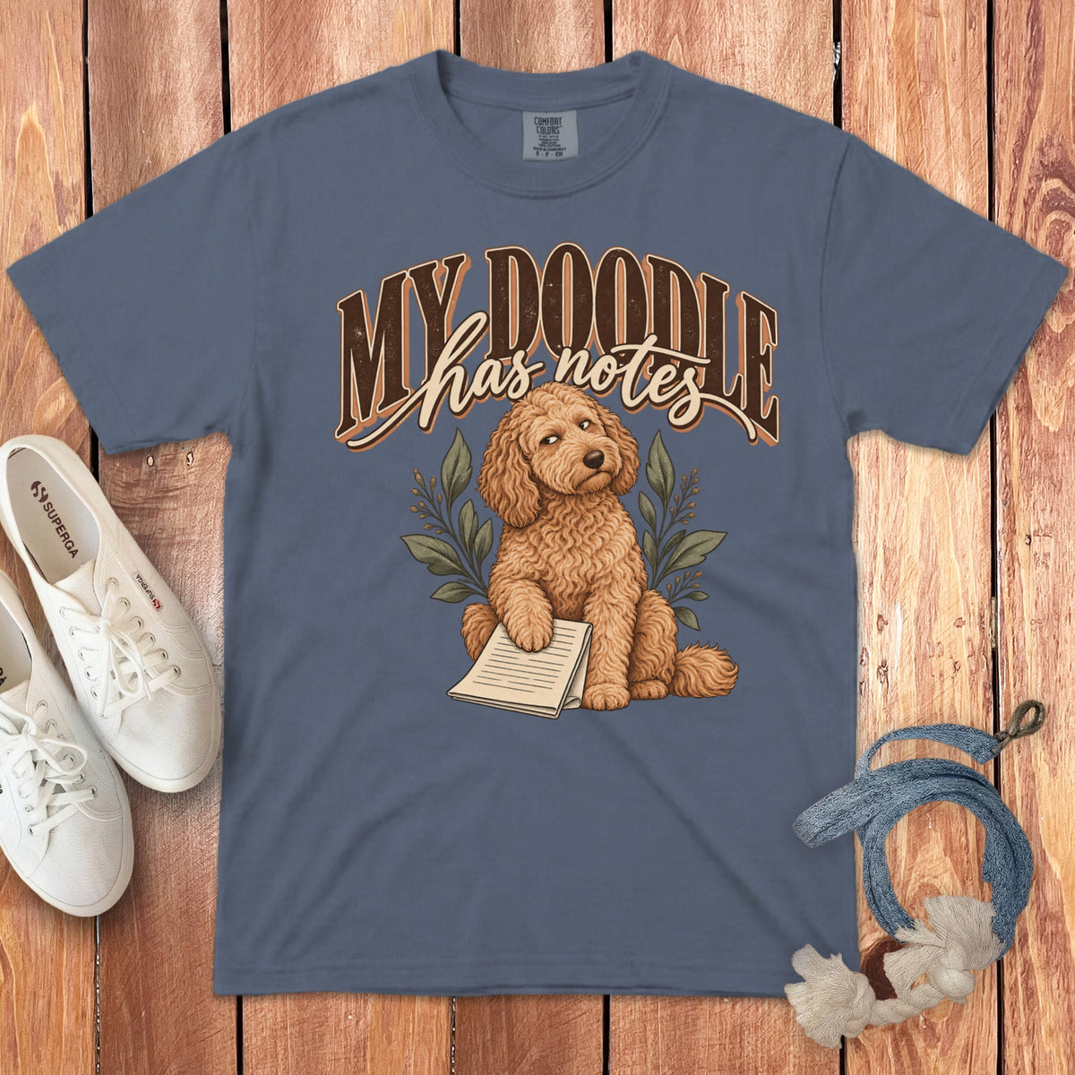 Slate blue t-shirt featuring a cartoon doodle dog holding notes, surrounded by white sneakers and a leash.