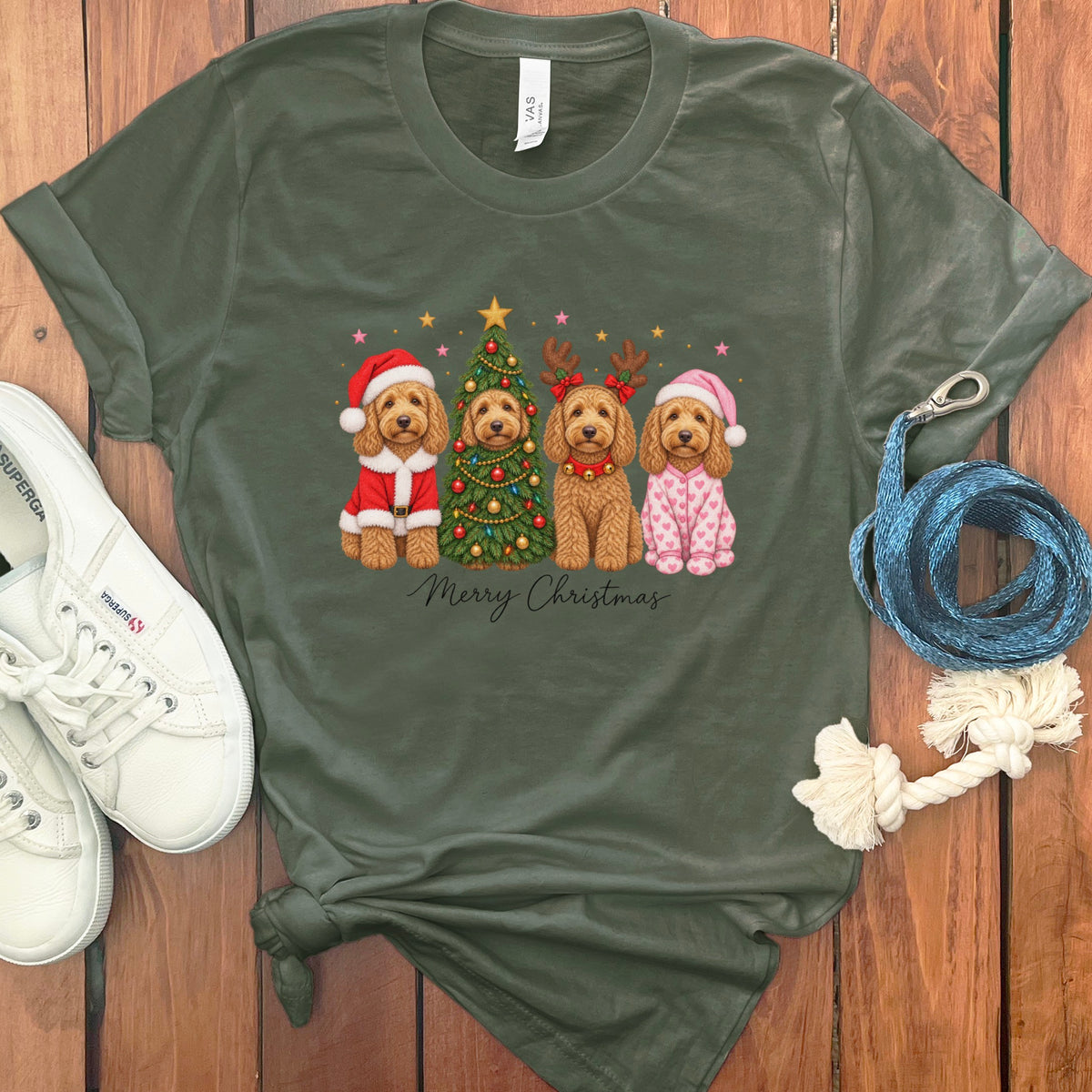 Dark green t-shirt with a graphic of four doodle dogs celebrating Christmas, surrounded by sneakers and a leash on a wooden surface.
