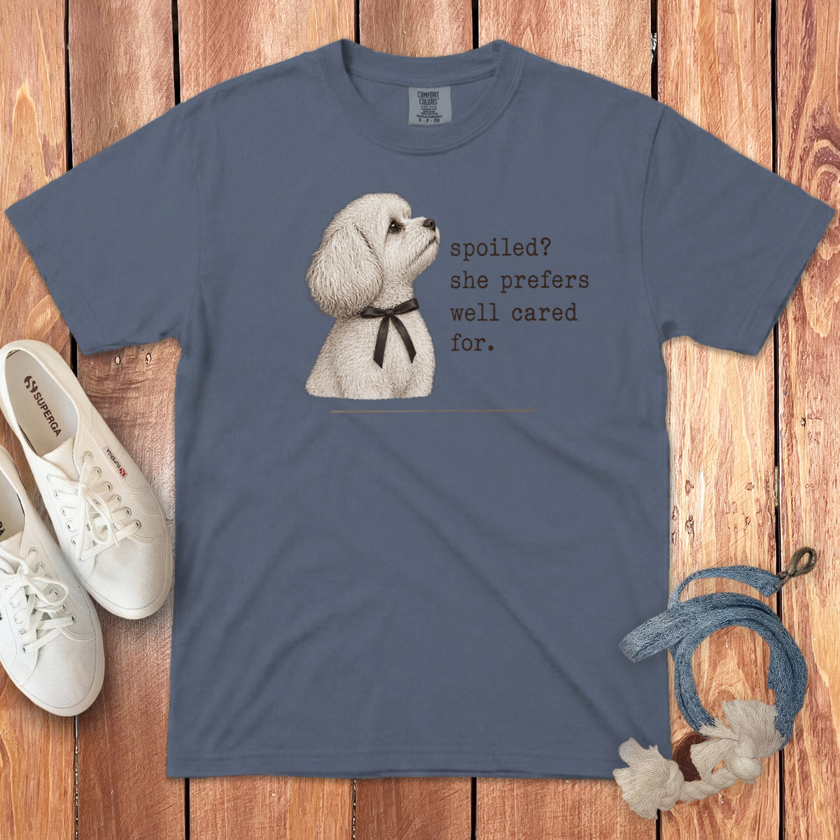 Blue t-shirt featuring a white poodle graphic and the text "spoiled? she prefers well cared for." laid flat on wood with white sneakers and a blue rope toy.