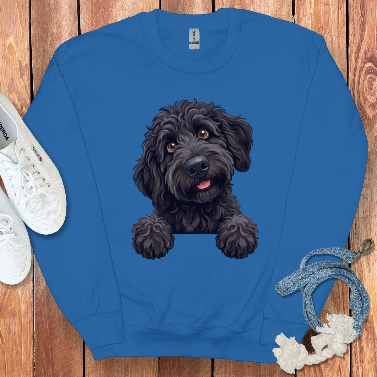 A blue crewneck sweatshirt featuring a graphic of a black poodle dog surrounded by sneakers and a leash on a wooden background.
