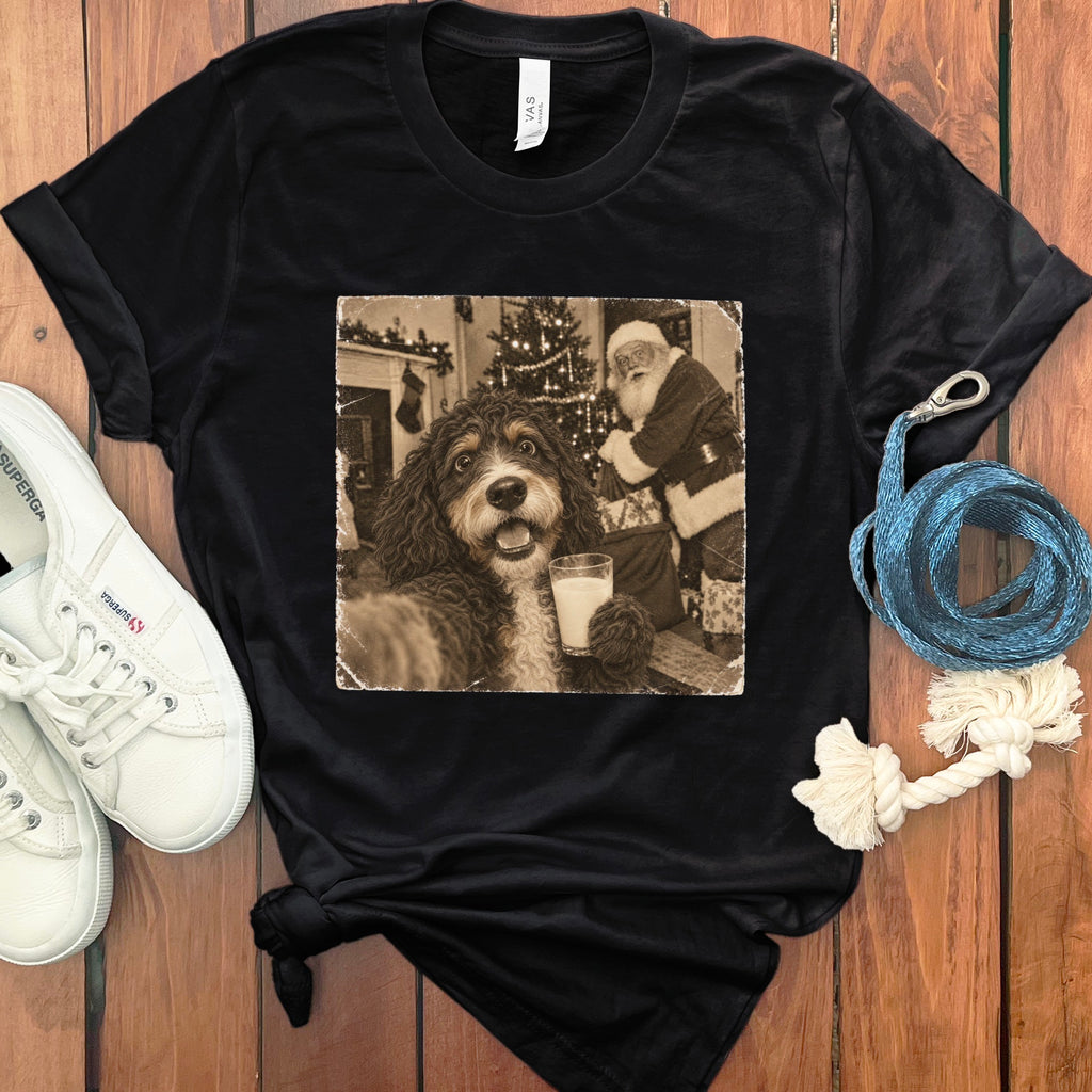 A black t-shirt features a graphic print of a dog holding a glass of milk with Santa Claus in the background, laid on a wooden surface beside white sneakers and a blue leash.