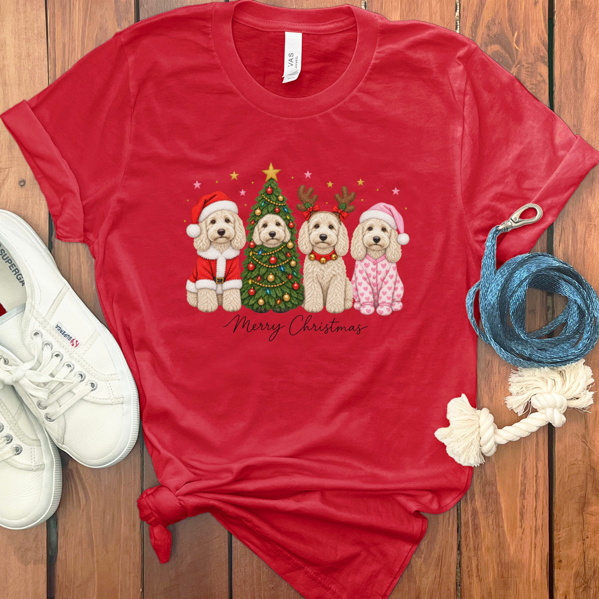 A red t-shirt with a graphic of four doodle dogs celebrating Christmas, surrounded by white sneakers and a blue leash.