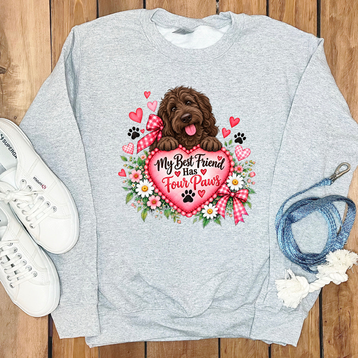 Light gray crewneck sweatshirt featuring a graphic of a brown dog resting on a pink heart with flowers and the text My Best Friend Has Four Paws, laid on wood with white sneakers and a blue leash.