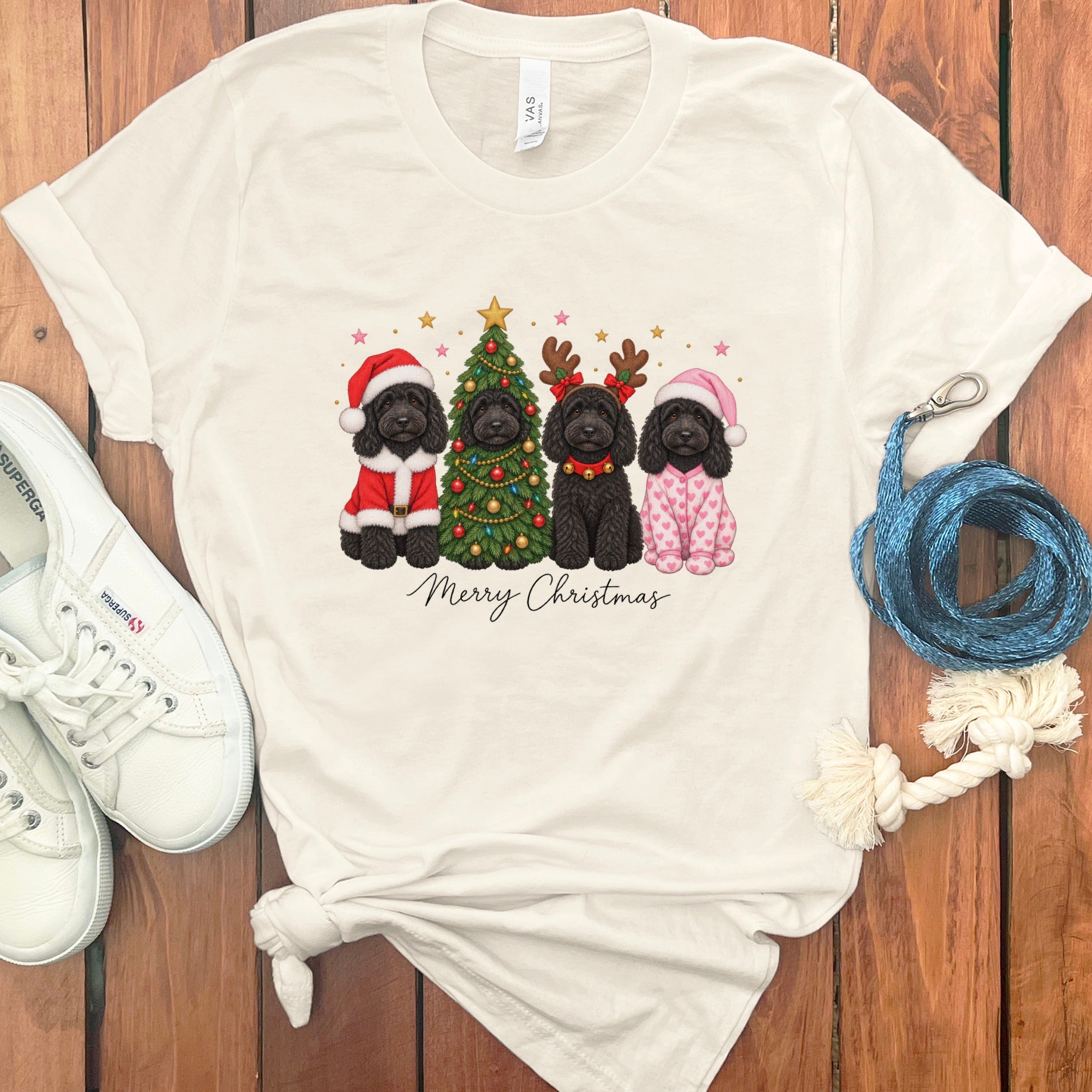 A cream-colored t-shirt with a graphic of four black doodle dogs celebrating Christmas, surrounded by white sneakers and accessories on a wooden background.