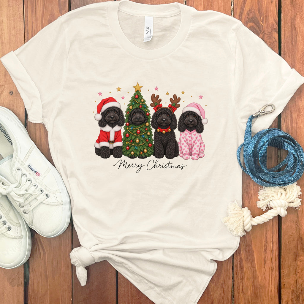 A cream-colored t-shirt with a graphic of four black doodle dogs celebrating Christmas, surrounded by white sneakers and accessories on a wooden background.
