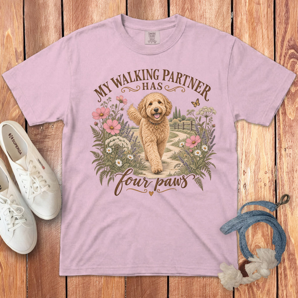 A pink t-shirt featuring a golden doodle dog graphic, text that reads My walking partner has four paws, and surrounded by flowers on a wooden background.
