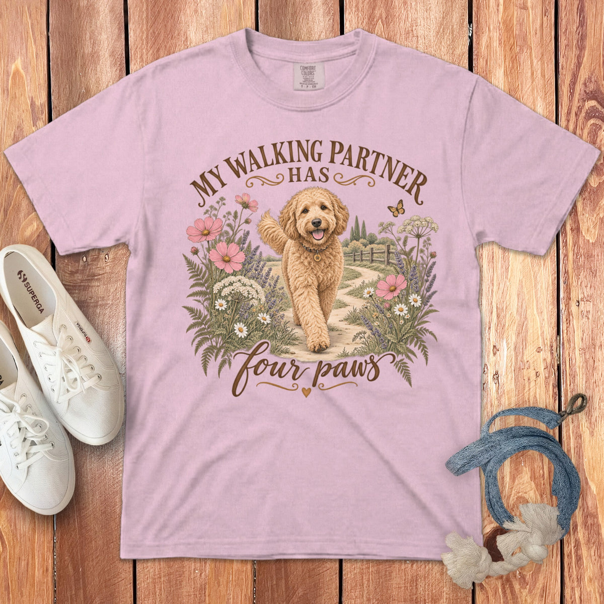 A pink t-shirt featuring a golden doodle dog graphic, text that reads My walking partner has four paws, and surrounded by flowers on a wooden background.
