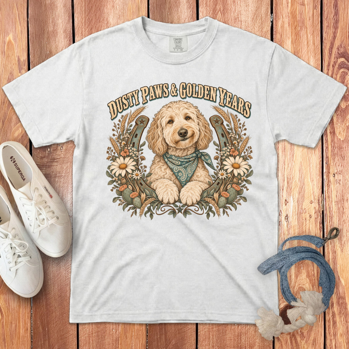 A white t-shirt featuring a graphic of a golden doodle wearing a bandana, surrounded by horseshoes and flowers, with the text DUSTY PAWS & GOLDEN YEARS.