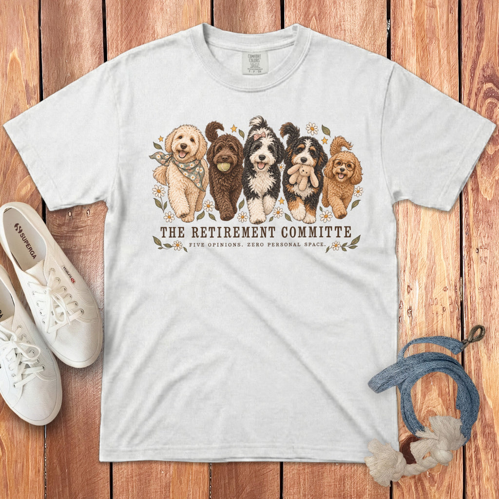 White t-shirt featuring a graphic of five doodle dogs and text reading The Retirement Committee on a wooden background.