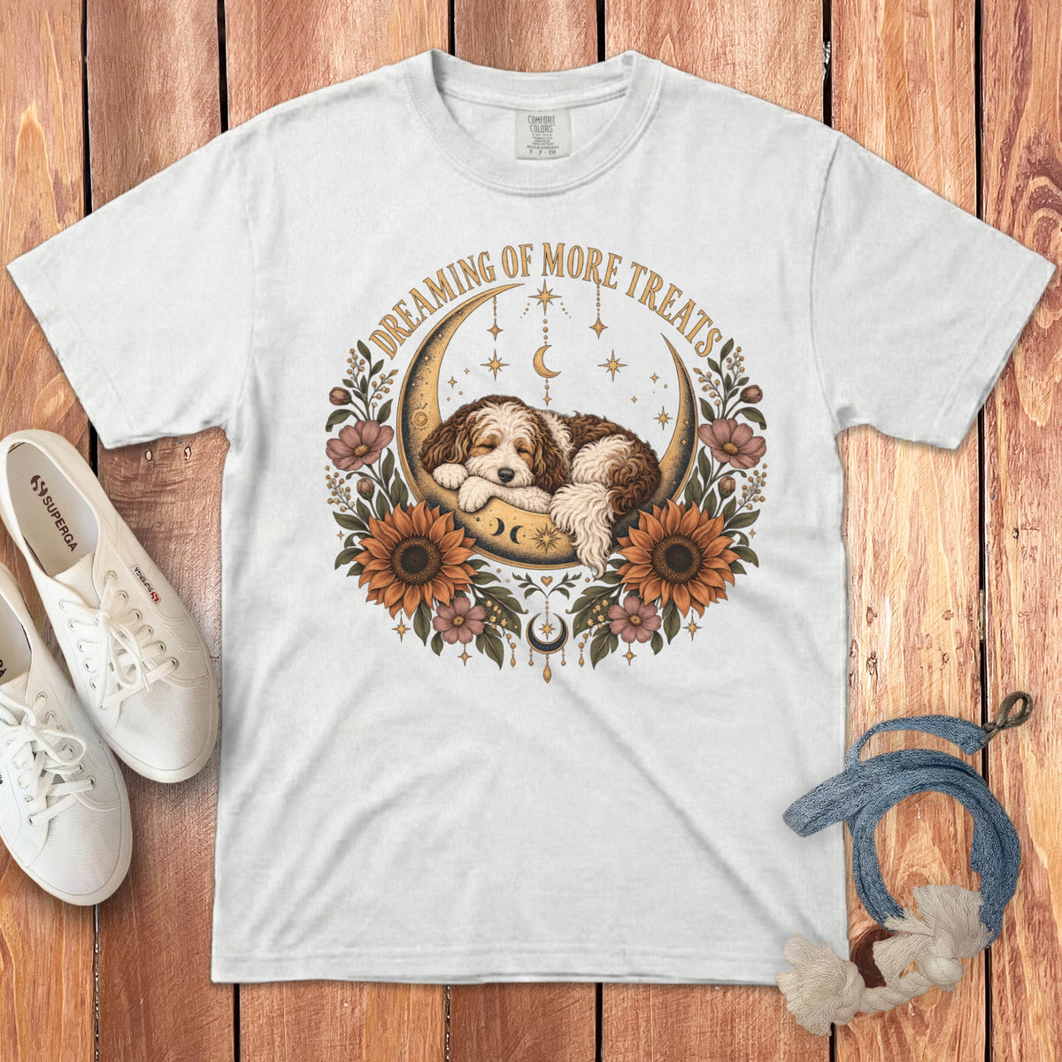 White t-shirt featuring a graphic of a puppy sleeping on a crescent moon surrounded by flowers, stars, and text reading Dreaming of More Treats.