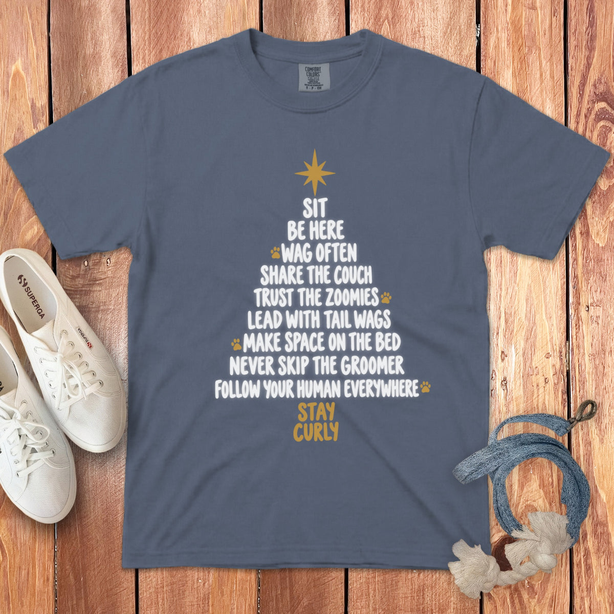 A slate blue crewneck t-shirt featuring a Christmas tree design made of dog-themed text on a wooden background with white sneakers and a rope toy.