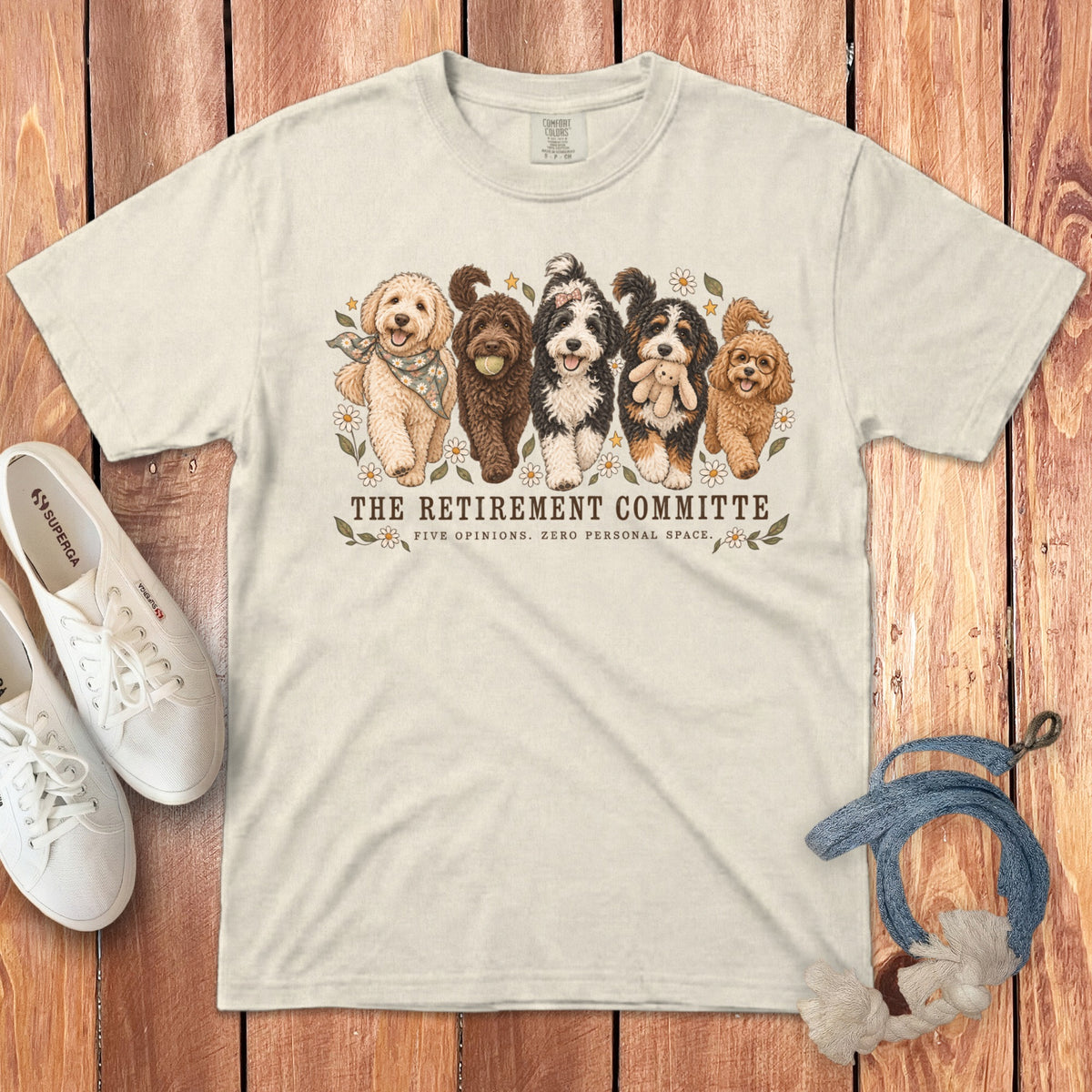A cream-colored t-shirt with a graphic of five dogs and text that reads The Retirement Committee, accompanied by white sneakers and a blue leash on a wooden background.
