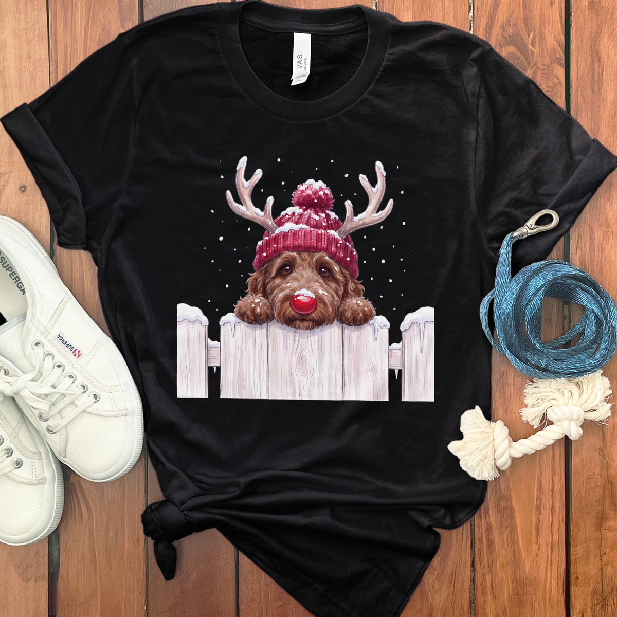 Black t-shirt with a brown dog wearing a beanie and antlers over a white fence.