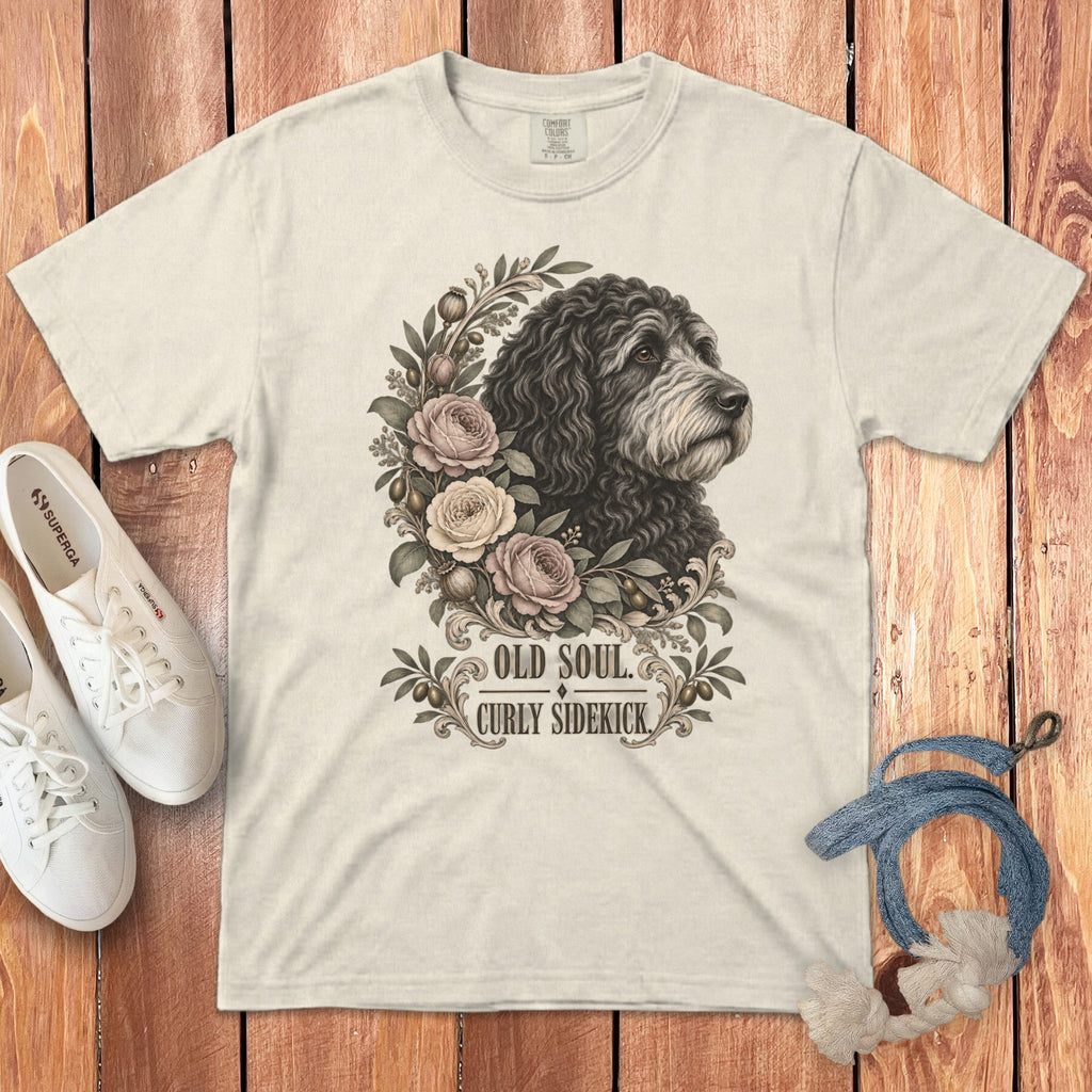 An off-white t-shirt featuring a black doodle dog surrounded by flowers and the text OLD SOUL CURLY SIDEKICK on a wooden background.