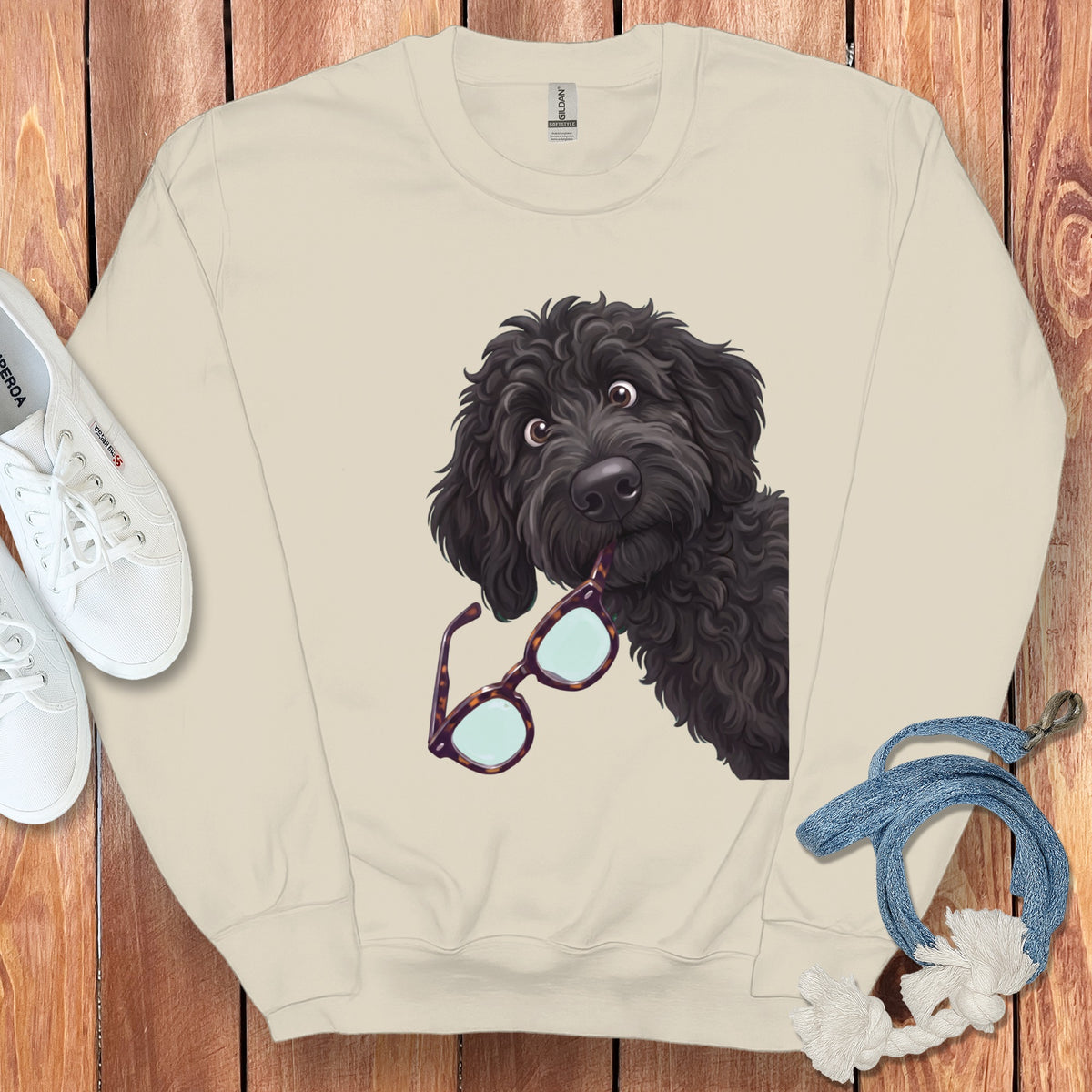 Beige crewneck sweatshirt with a graphic of a black poodle holding glasses in its mouth, laid flat on a wooden surface.