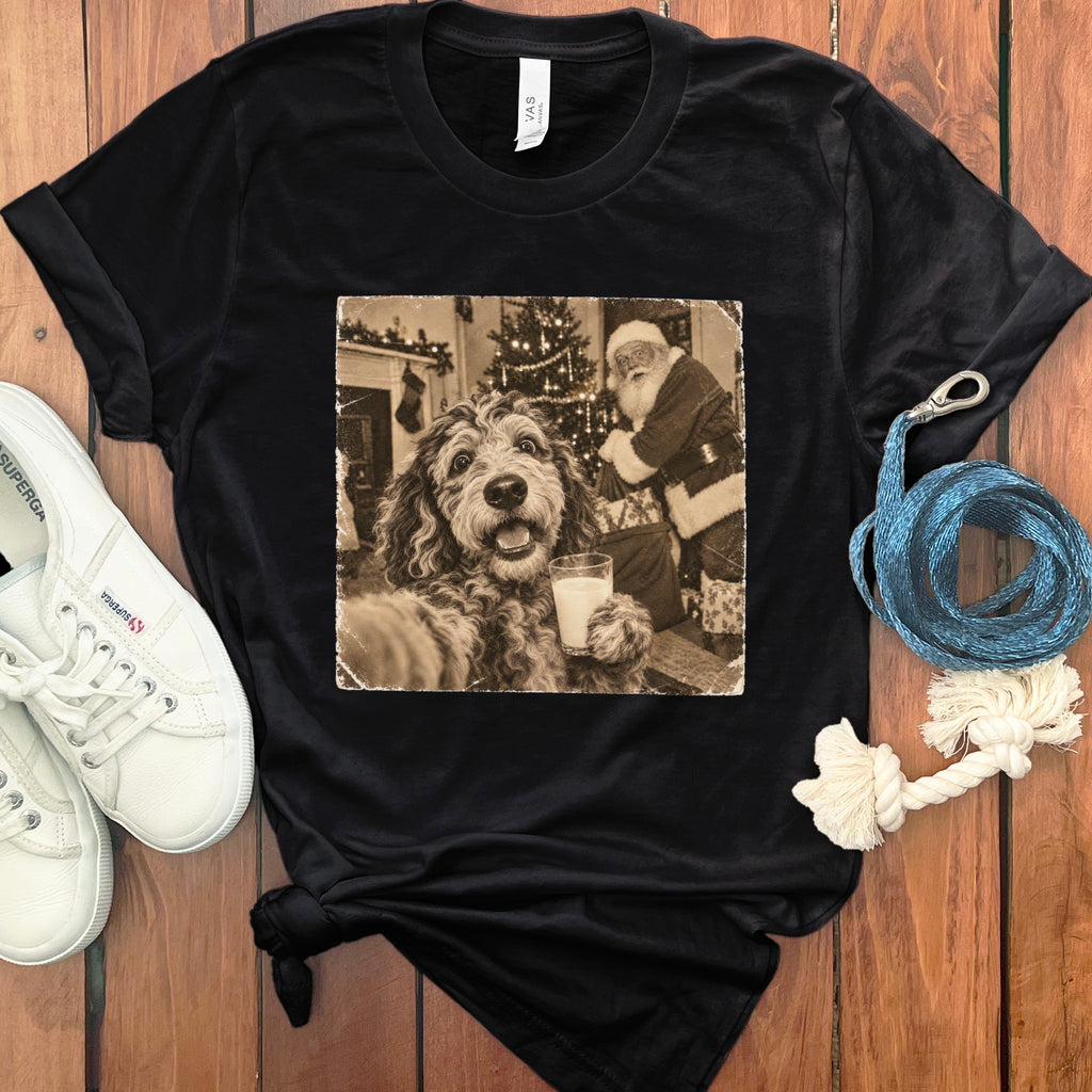 A black t-shirt printed with a sepia-toned graphic of a dog holding a glass of milk in front of Santa Claus.