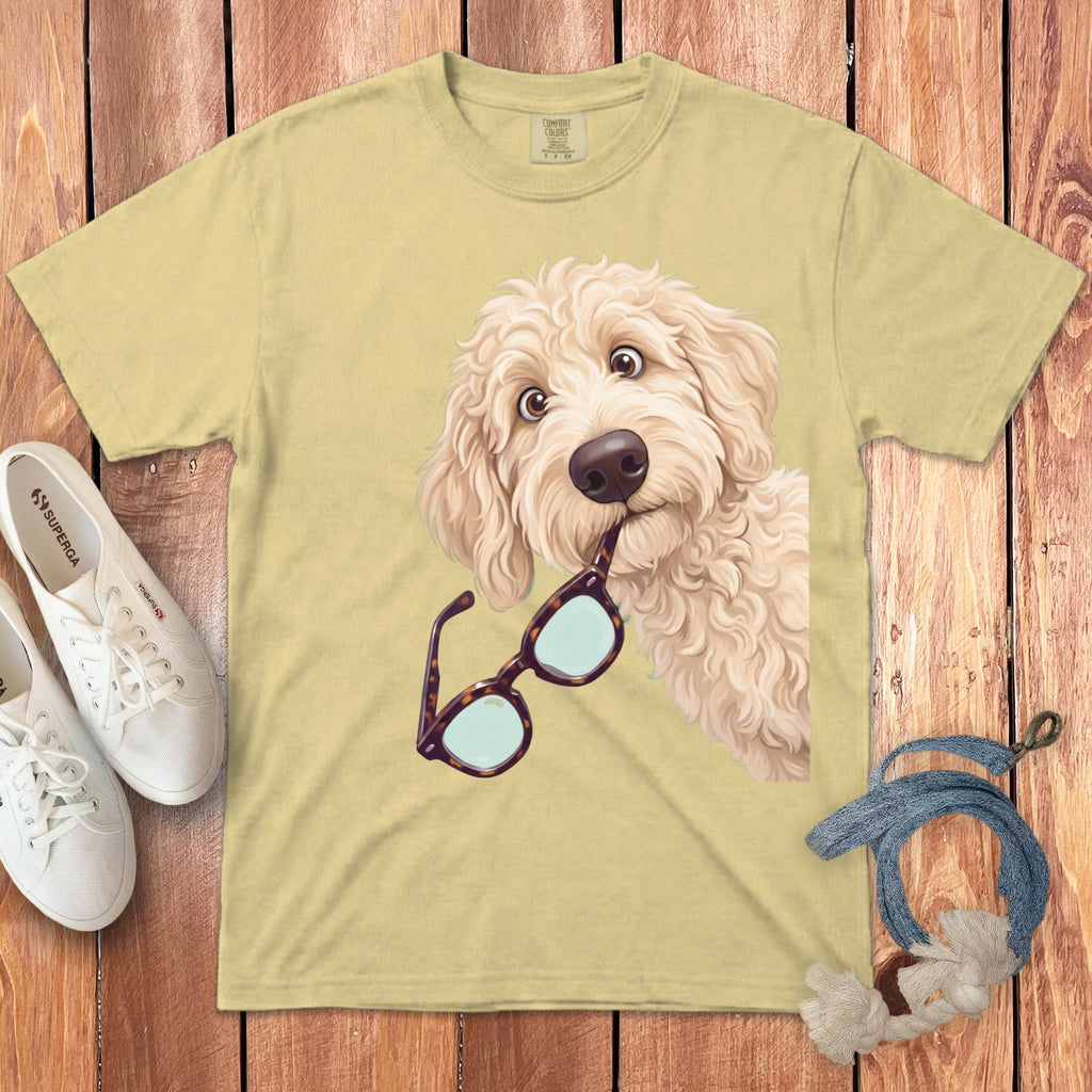 Light yellow t-shirt featuring a doodle dog holding glasses in its mouth, laid on wood.