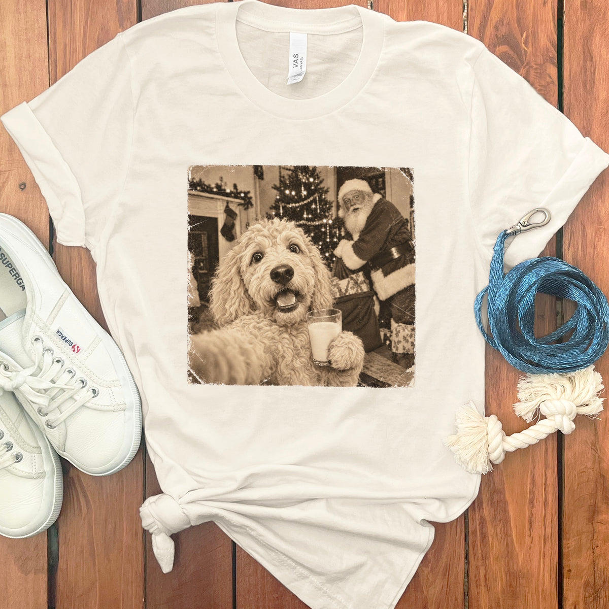 White t-shirt featuring a vintage graphic of a fluffy dog holding a glass of milk with Santa Claus in the background, laid on a wooden surface with white sneakers and a blue leash.