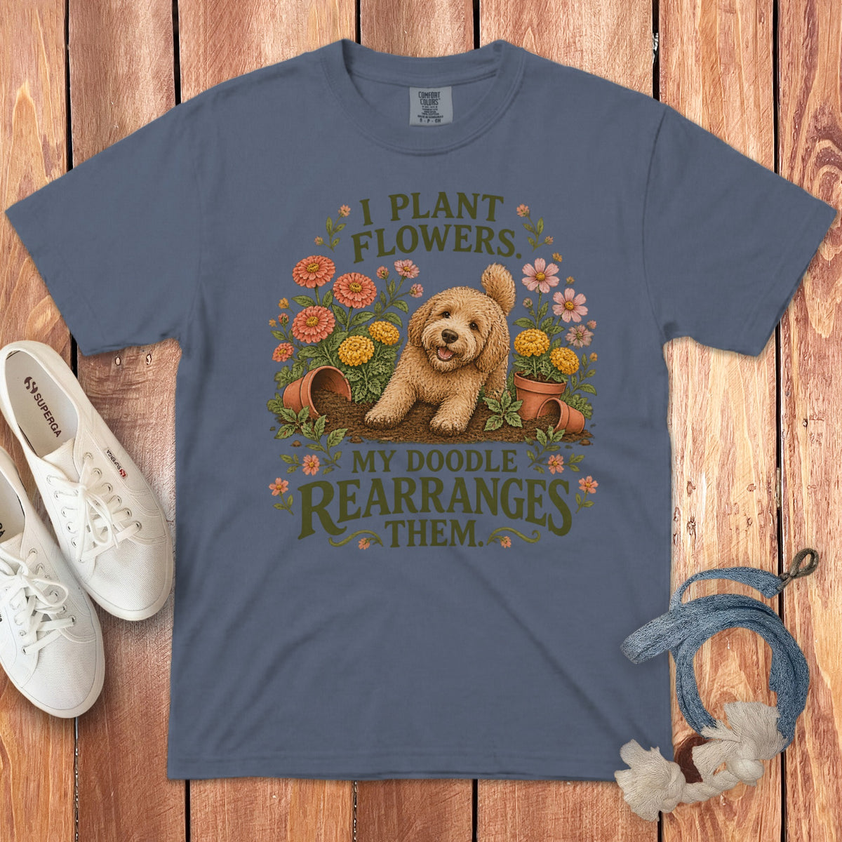 Slate blue t-shirt featuring a playful doodle dog among flowers and text reading I plant flowers my doodle rearranges them.