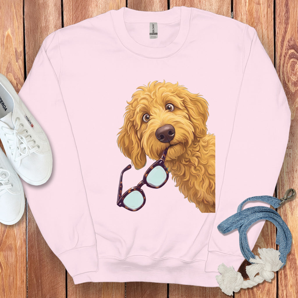 Light pink crewneck sweatshirt with a golden doodle dog graphic holding glasses in its mouth, laid flat on wood.
