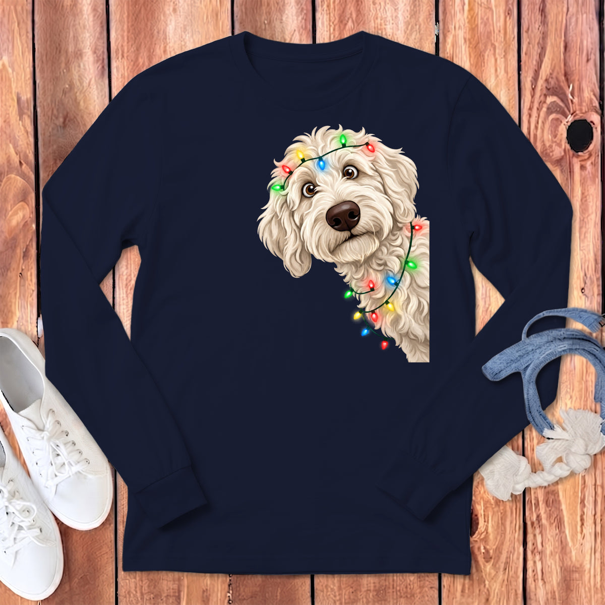 Cream doodle dog peeking from behind colorful Christmas lights on navy long-sleeve shirt.