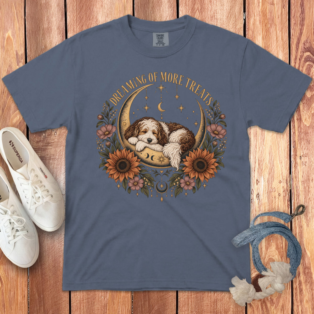 A blue t-shirt featuring a sleeping puppy in a crescent moon surrounded by flowers and the text Dreaming of More Treats.