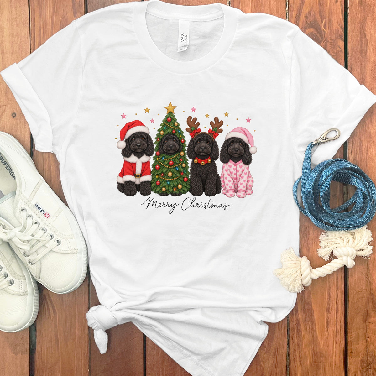 A white t-shirt featuring a graphic of four black doodle dogs celebrating Christmas, surrounded by white shoes and a blue leash on a wooden background.
