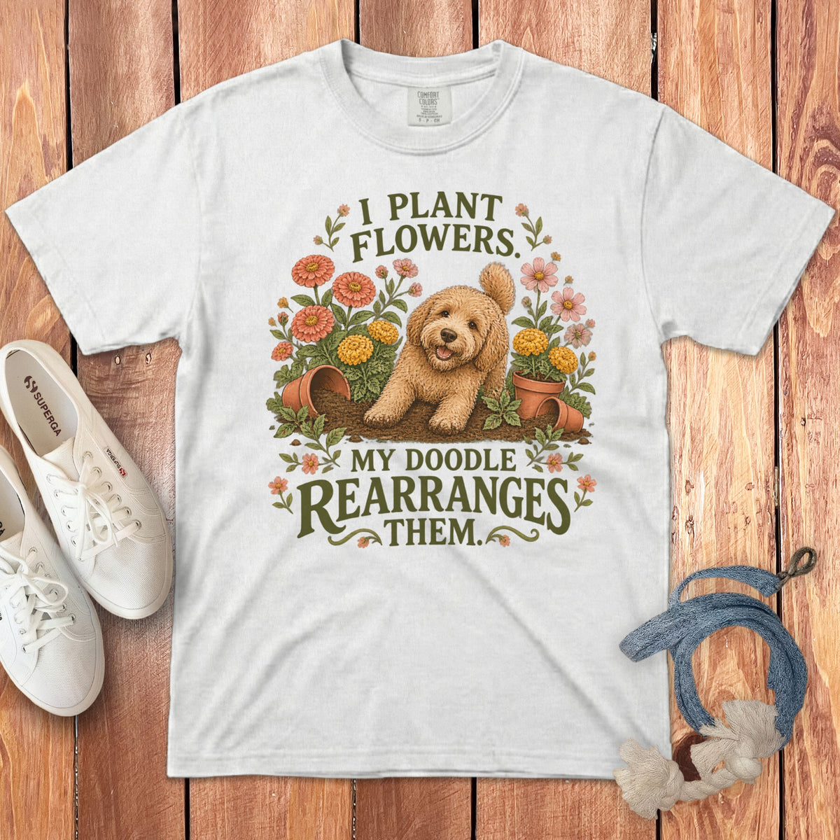 White t-shirt featuring a graphic of a Goldendoodle digging in a flower garden and text that reads I plant flowers, my doodle rearranges them.