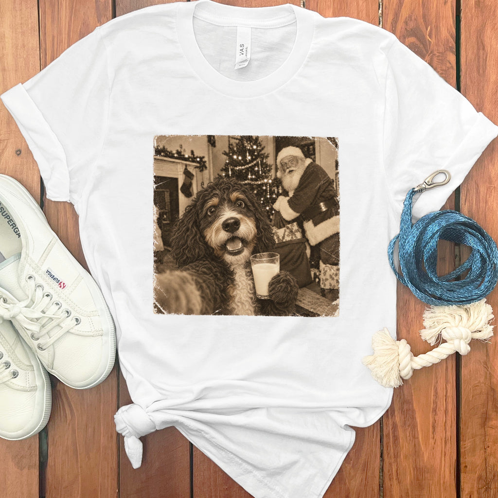 A white t-shirt featuring a retro graphic of a dog holding a glass of milk with Santa Claus in the background, laid on a wooden surface with white sneakers and a blue leash.