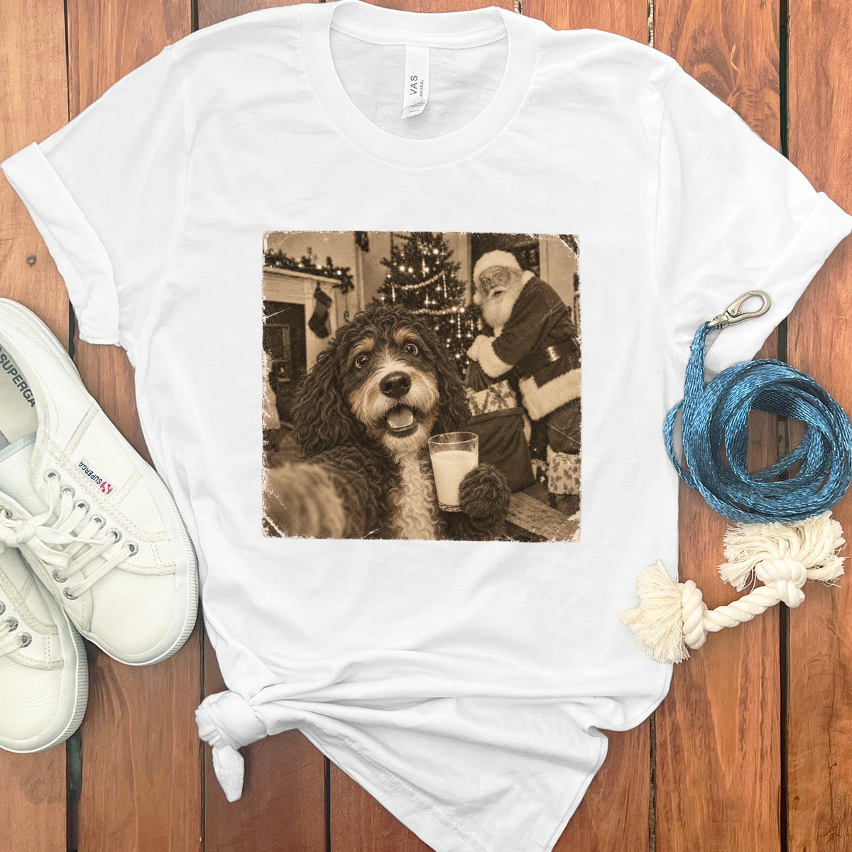 A white t-shirt featuring a retro graphic of a dog holding a glass of milk with Santa Claus in the background, laid on a wooden surface with white sneakers and a blue leash.