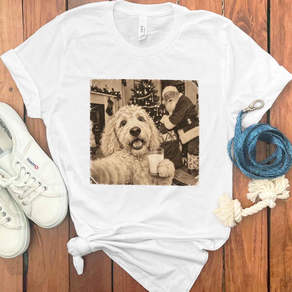 A white t-shirt featuring a sepia-toned graphic of a fluffy dog taking a selfie with Santa Claus.