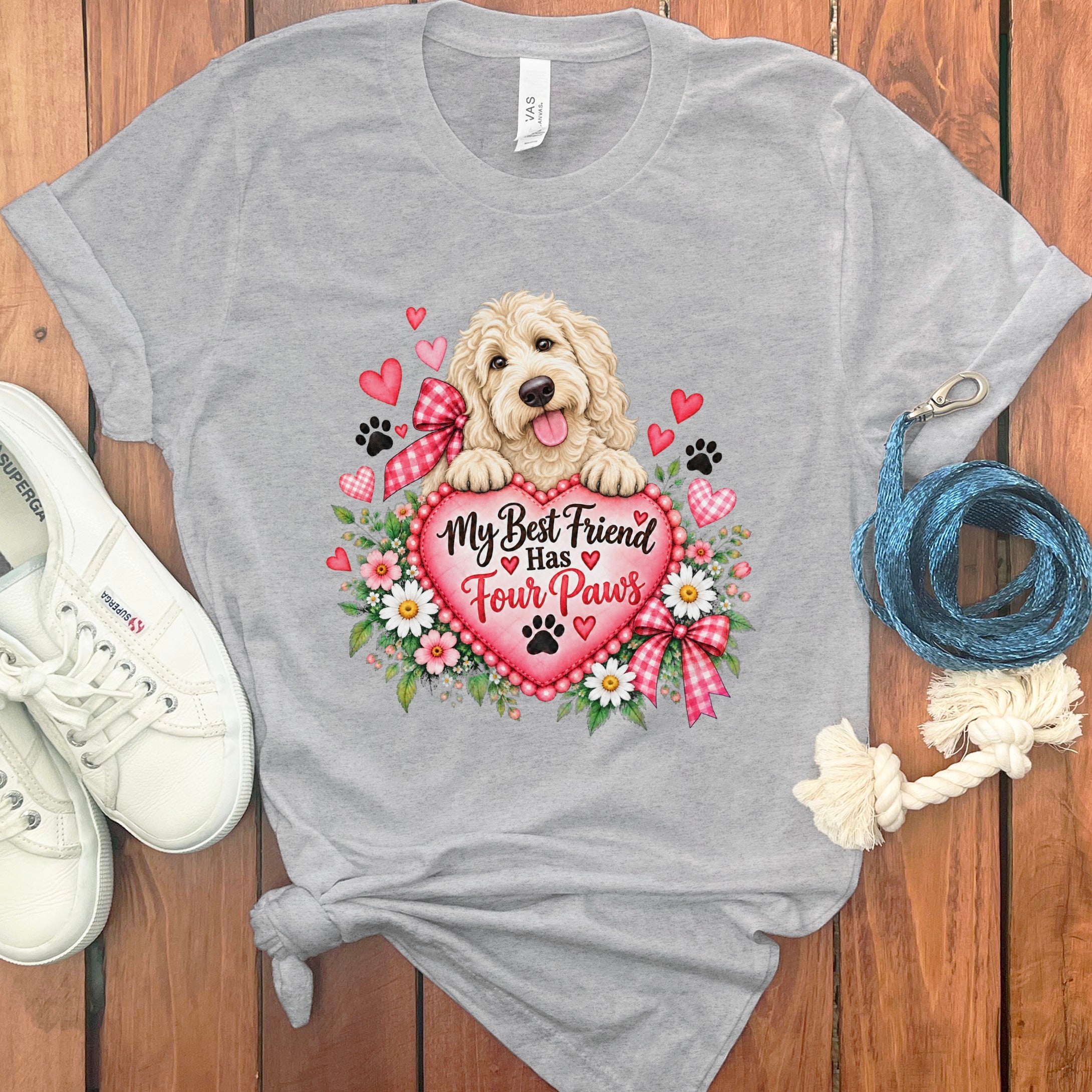 Heather gray crewneck t-shirt featuring a graphic of a fluffy dog holding a large heart, surrounded by flowers, hearts, and paw prints, flanked by white sneakers and a blue leash on a wooden background.