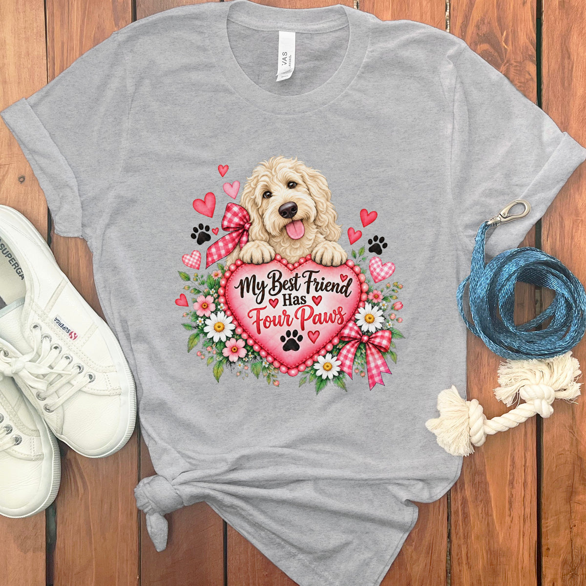 Heather gray crewneck t-shirt featuring a graphic of a fluffy dog holding a large heart, surrounded by flowers, hearts, and paw prints, flanked by white sneakers and a blue leash on a wooden background.