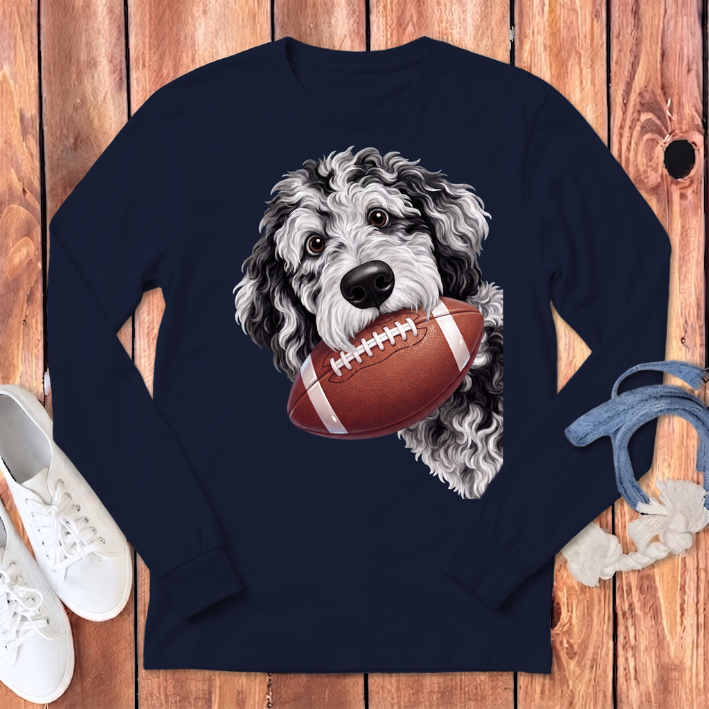 Black and white dog with curly fur holds a football in its mouth on a navy long-sleeve shirt.