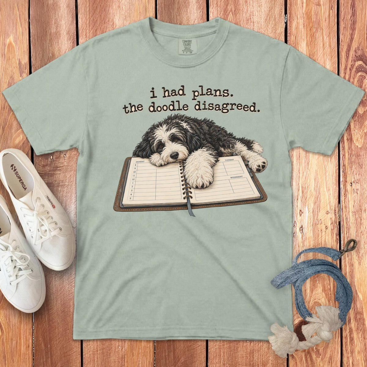 Sage green t-shirt with a graphic of a dog lying on a planner and text that reads i had plans. the doodle disagreed.