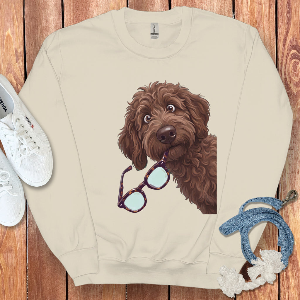 Cream crewneck sweatshirt featuring a cartoon brown poodle holding glasses in its mouth, surrounded by white sneakers and a blue rope on a wooden background.