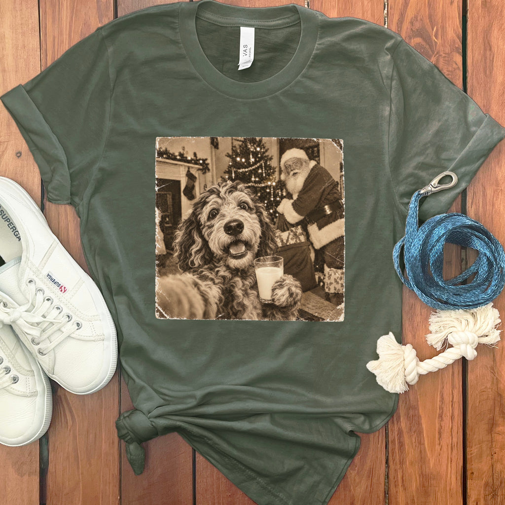 Olive green t-shirt featuring a retro graphic of a dog holding a glass of milk near Santa Claus, arranged on a wooden surface with white sneakers and a blue leash.