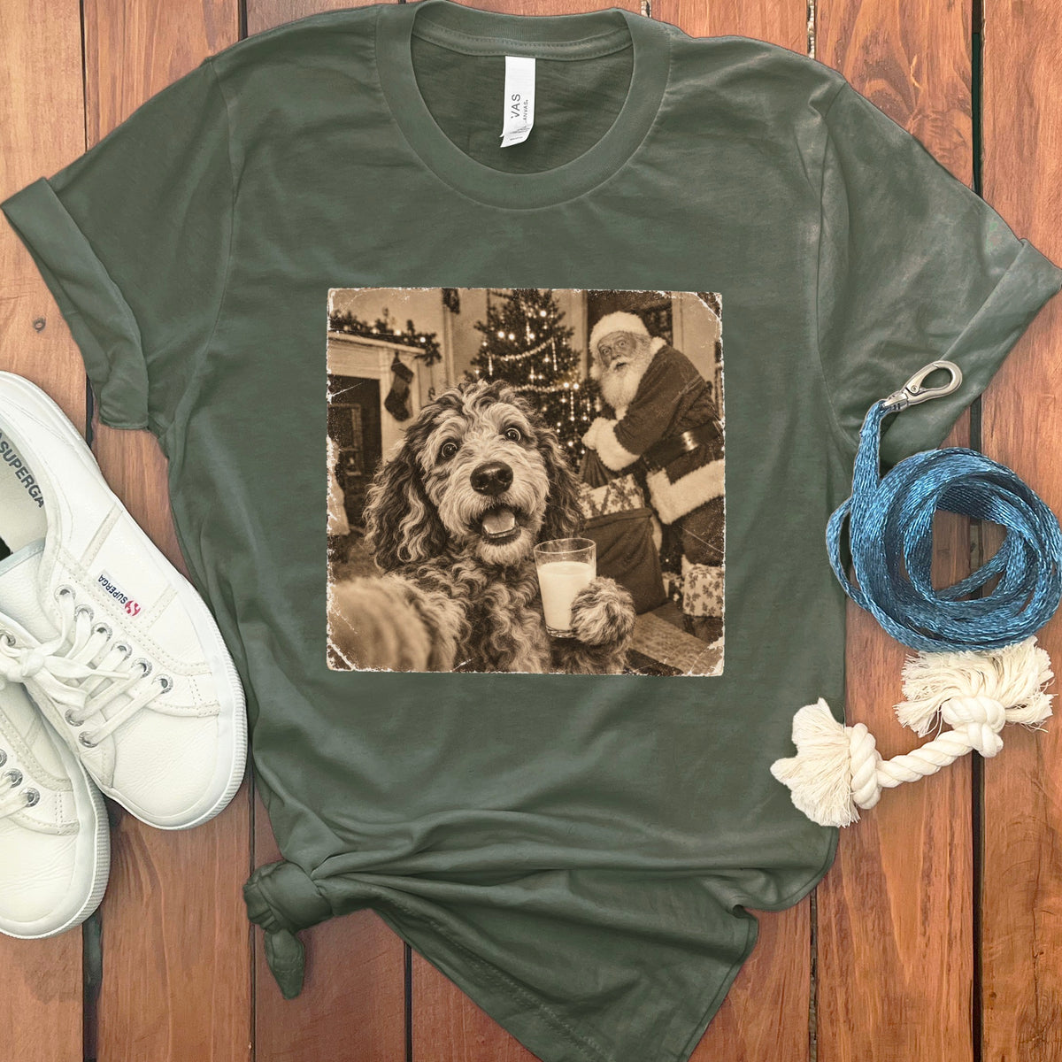 Olive green t-shirt featuring a retro graphic of a dog holding a glass of milk near Santa Claus, arranged on a wooden surface with white sneakers and a blue leash.