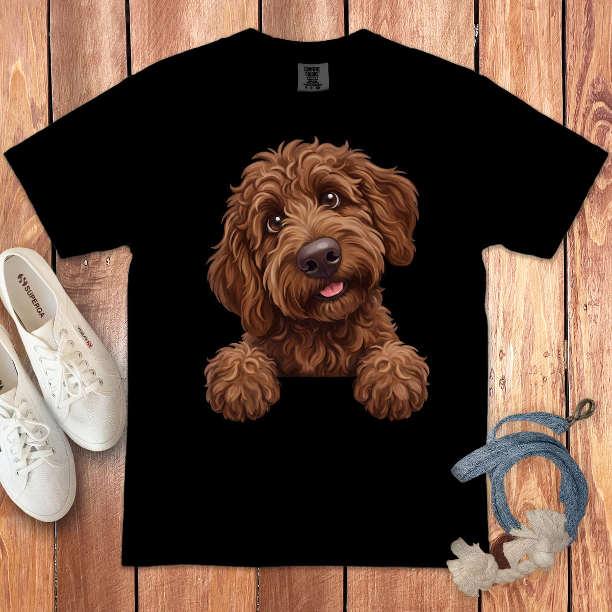 A black t-shirt printed with a cute brown doodle dog graphic, surrounded by white sneakers and a rope toy on a wooden background.