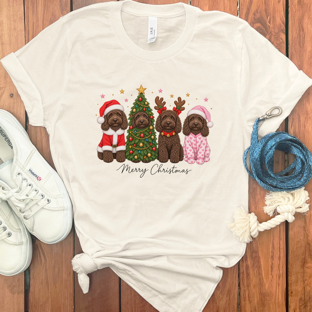 A cream-colored t-shirt featuring a graphic of four doodle dogs celebrating Christmas, surrounded by sneakers and accessories on a wooden surface.