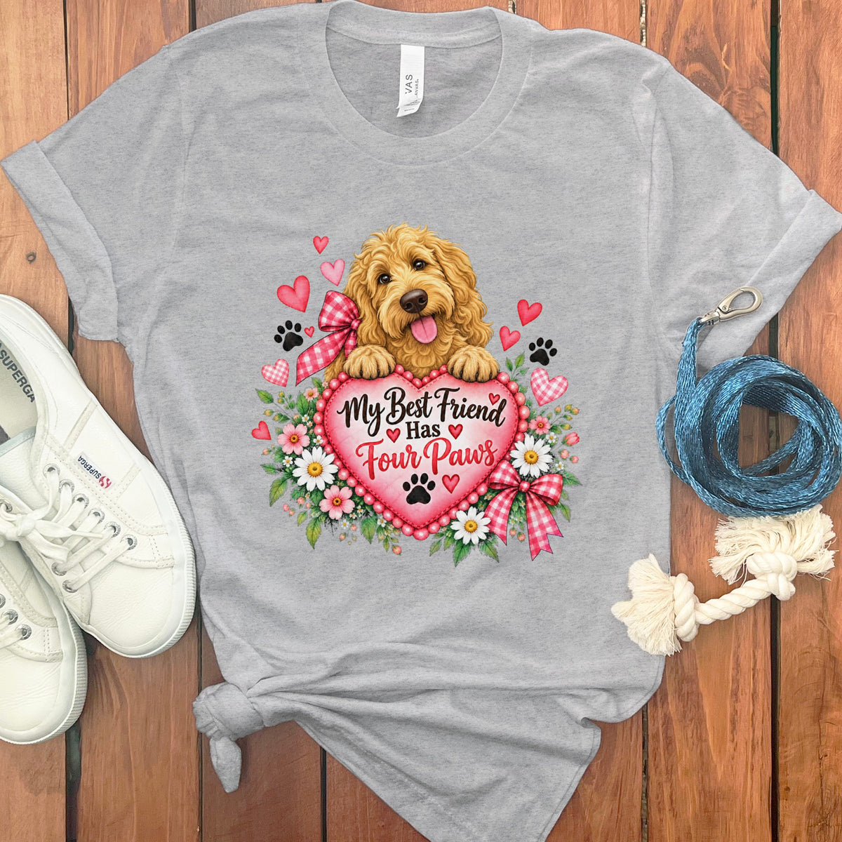A gray t-shirt featuring a graphic of a golden doodle holding a pink heart that says My Best Friend Has Four Paws, with white sneakers and a blue leash on a wood background.