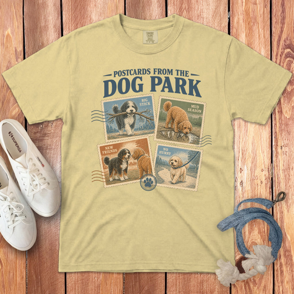 Yellow t-shirt featuring Postcards from the Dog Park graphic with four illustrated dog stamps on a wooden background.