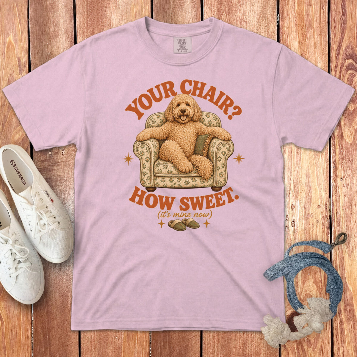 A light pink t-shirt featuring a graphic of a fluffy dog sitting in an armchair with text that reads Your Chair? How Sweet.