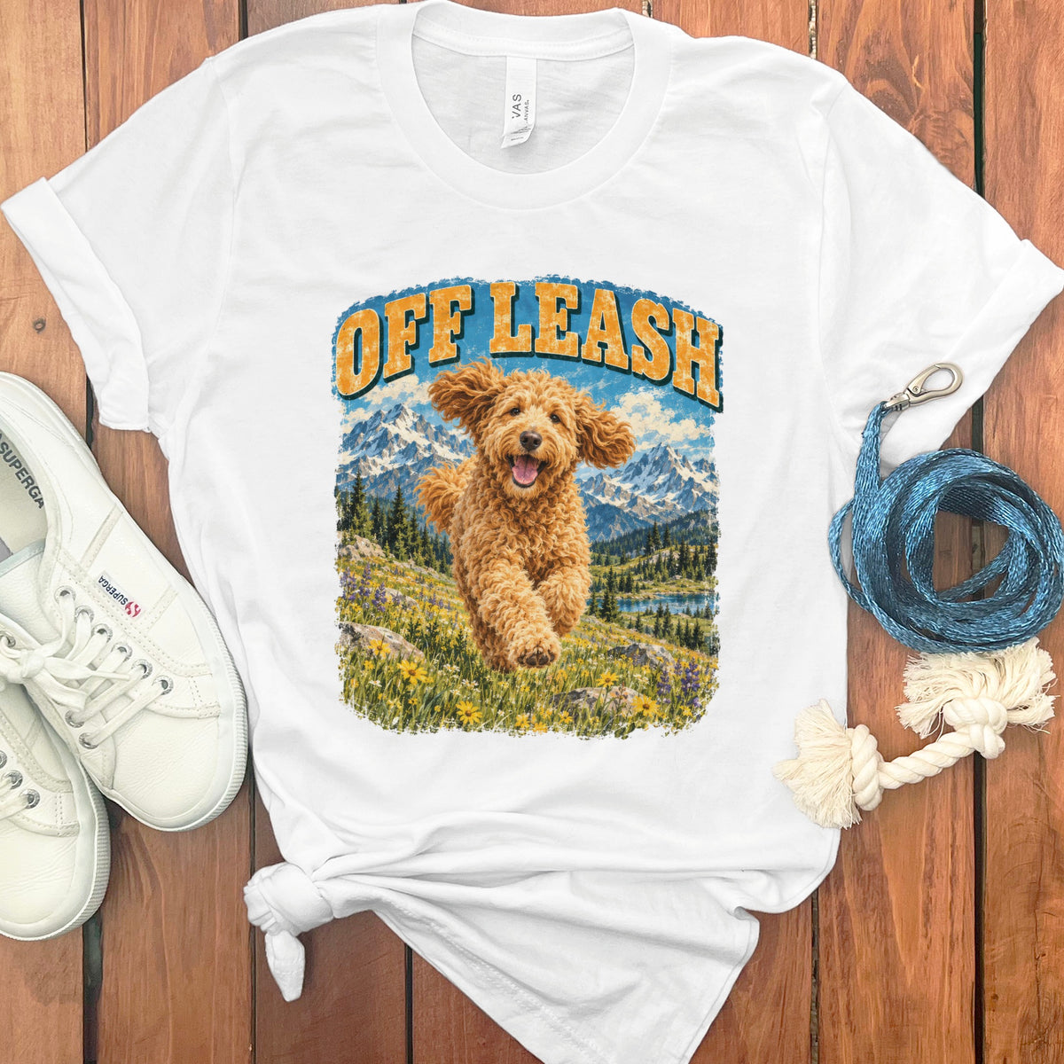 White t-shirt with "Off Leash" text and running golden doodle in mountain landscape.