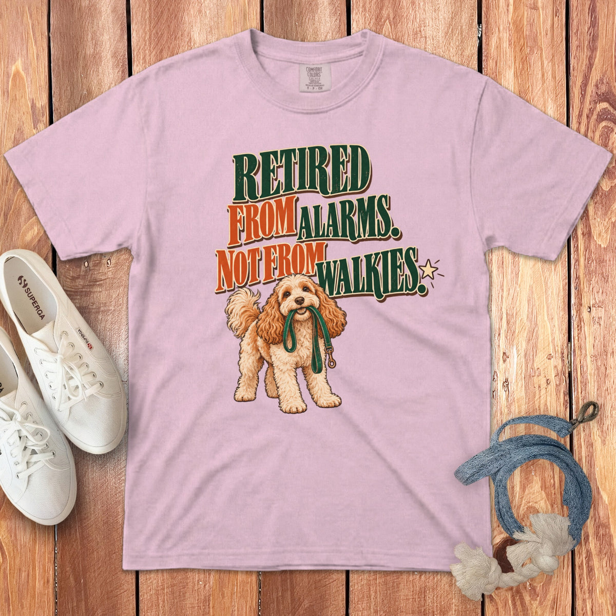 A light pink t-shirt featuring the text "RETIRED FROM ALARMS NOT FROM WALKIES" alongside a drawing of a fluffy dog holding a leash in its mouth.