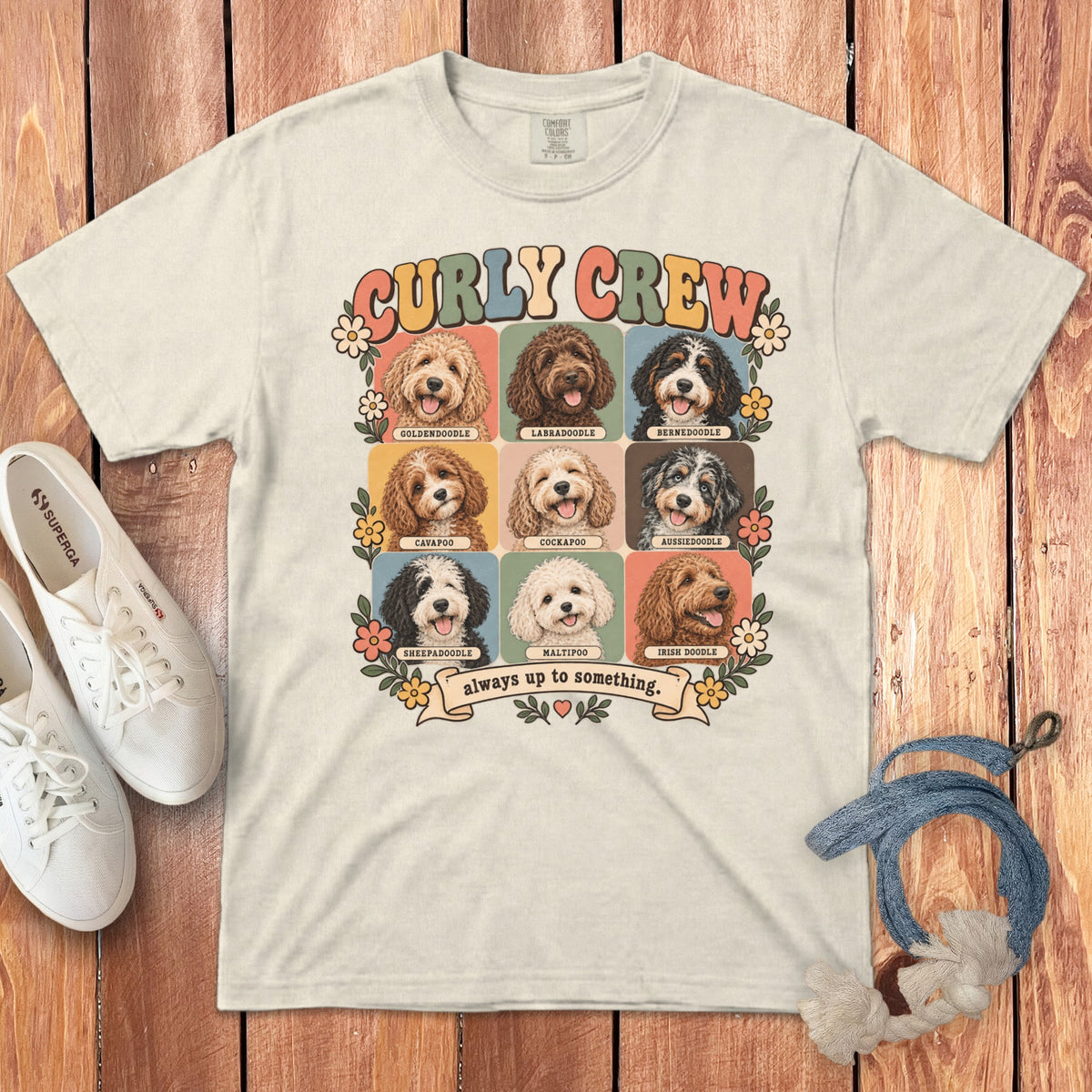 Beige t-shirt featuring a retro graphic of nine doodle dog breeds, text reading Curly Crew, and flowers on a wooden background with white sneakers and a leash.